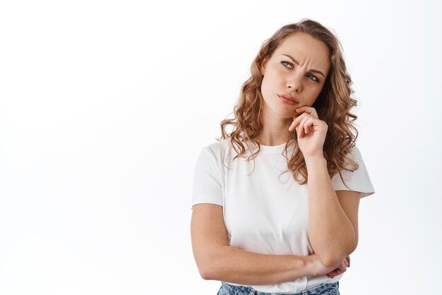 Thoughtful blond woman pondering frowning and looking puzzled at upper left corner promo text thinking making decision standing over white background