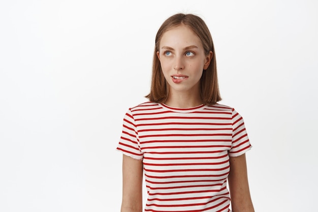 Free photo thoughtful blond woman wants something, biting lip and looking aside with tempted look, has desire, wants to try something tasty, standing against white wall.