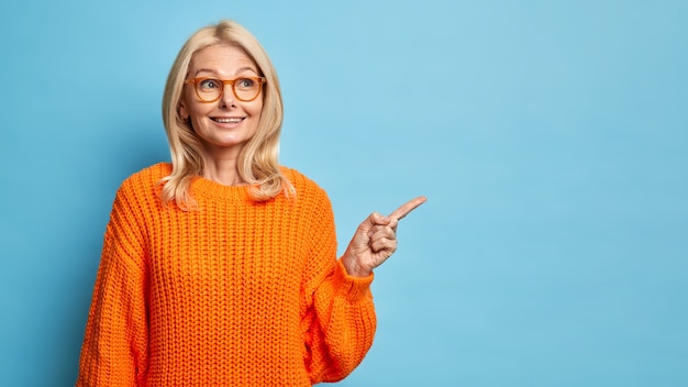 Thoughtful blonde forty years old European woman wears spectacles and knitted orange sweater pointing at copy space
