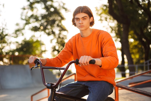 Вдумчивый мальчик в оранжевом пуловере сидит на велосипеде, мечтательно глядя в камеру, проводя время в современном скейтпарке