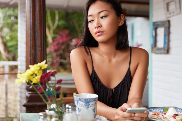 Thoughtful brunette Asian female in casual clothing uses modern cell phone for messaging with friends, spends free time in coffee shop, enjoys tasty dish and hot drink, looks pensively down.
