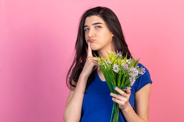 Thoughtful brunette holding bunch of flowers and thinking on pink background High quality photo