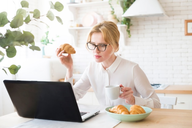 Вдумчивый бизнес женщина, используя ноутбук во время еды круассан