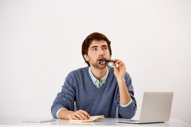 Thoughtful businessman sitting at the office desk, biting glasses rim, look up thinking, search inspiration as writing in notebook