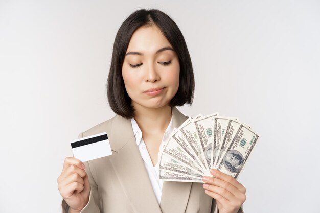 Thoughtful businesswoman korean corporate woman showing credit card and money cash dollars in hands standing over white background and thinking