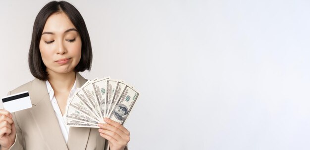 白い背景の上に立って考えて手にクレジットカードとお金の現金ドルを示す思いやりのある実業家韓国企業の女性