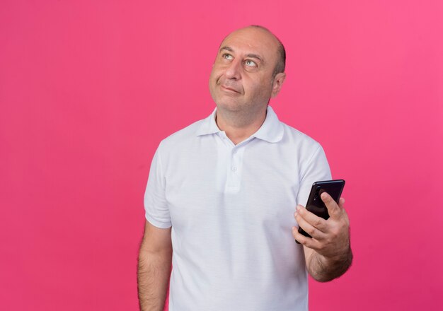 Thoughtful casual mature businessman looking up and holding mobile phone isolated on pink background with copy space