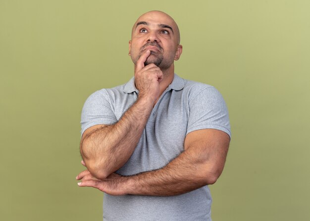 Thoughtful casual middle-aged man keeping finger on chin looking up isolated on olive green wall