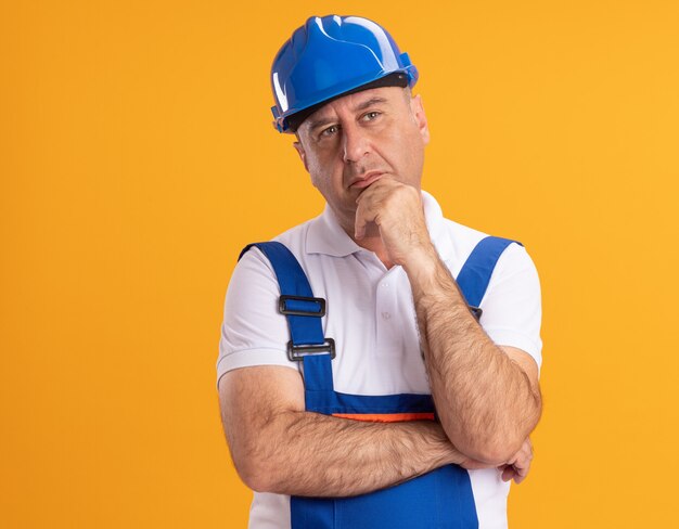 Вдумчивый кавказский взрослый строитель в униформе держит подбородок, глядя в сторону на оранжевом