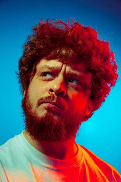 Thoughtful. Caucasian close up man's portrait isolated on blue  wall in red neon light. Beautiful male model, red curly hair. Concept of human emotions, facial expression, sales, ad.