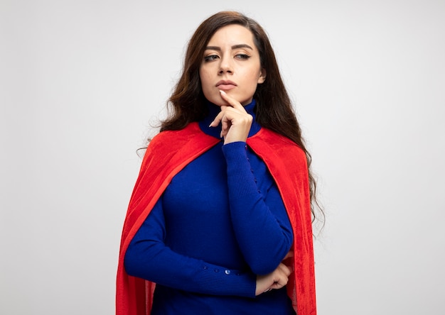 Thoughtful caucasian superhero girl with red cape puts hand on chin looking at side isolated on white wall with copy space