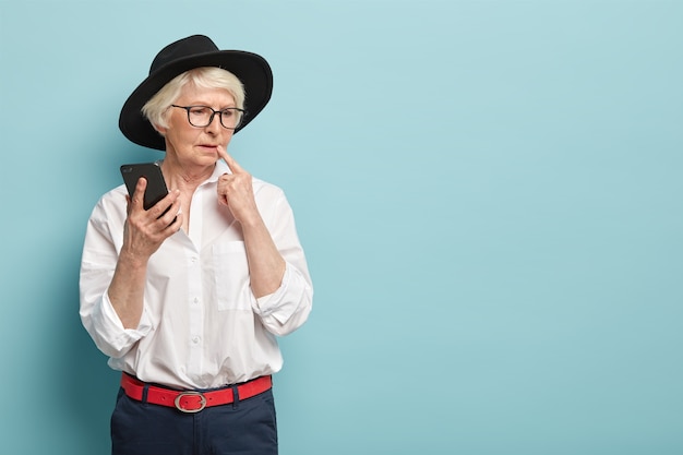 Free photo thoughtful charismatic senior lady dressed in fashionable outfit, thinks about message content, holds modern cell phone