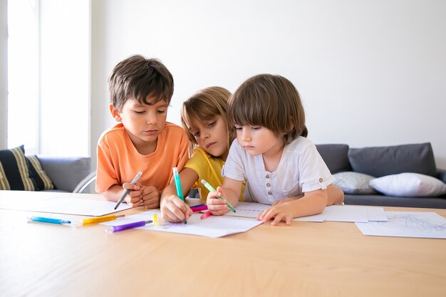 Thoughtful children painting with markers in living room. Caucasian lovely boys and blonde girl sitting at table, drawing on paper and playing together. Childhood, creativity and weekend concept