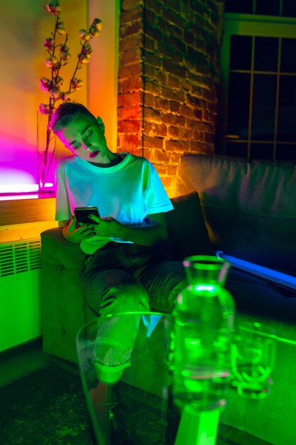 Thoughtful. Cinematic portrait of stylish woman in neon lighted interior. Toned like cinema effects, bright neoned colors. Caucasian model using smartphone in colorful lights indoors. Youth culture.