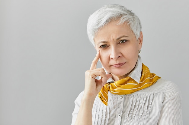 Thoughtful concentrated senior woman with gray pixie hair having memory problems, trying to recollect something, touching face. Serious mature lady posing with deep in thoughts pensive look