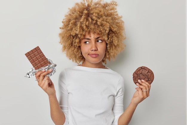 思いやりのある混乱した女性はチョコレートとクッキーのどちらかを選択しますカジュアルなセーターに身を包んだ甘い歯はダイエットを続けます白い背景の上に隔離されたおいしい有害な食べ物を食べたいという誘惑を感じます