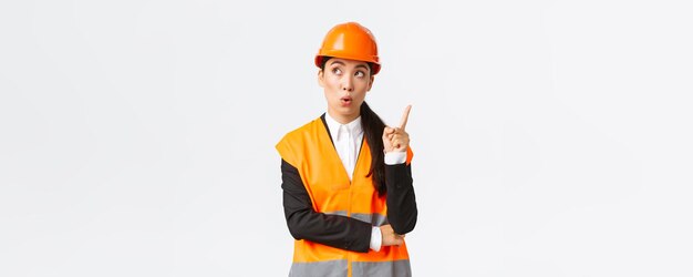 Thoughtful creative female asian engineer architect in safety helmet and reflective jacket raising index finger eureka gesture have interesting idea suggest plan have solution white background