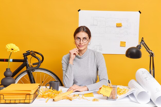 Thoughtful creative female worker dreams about holidays while working in office develops new business project makes blueprints wears spectacles poses in coworking space analyzes information.