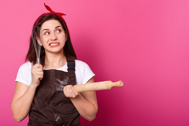 Thoughtful creative worker touching face with whisk, holding rolling pin in one hand, looking aside, wearing brown dirty apron