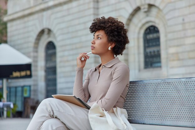 Thoughtful curly female student looks away makes notes in notepad holds pen and notebook sits on bench dressed in casual jumper and trousers writes information to textbook Studying outdoors