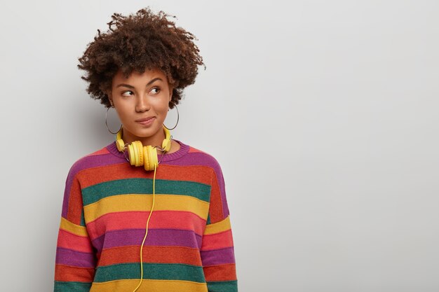 thoughtful curly haired woman looks aside with pensive expression, wears headphones and striped jumper, has plan in mind, poses over white background