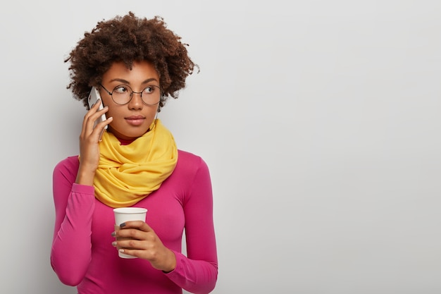 思いやりのある巻き毛の女性は、携帯電話を介して電話で話をし、コーヒーを飲み、会話を楽しんで、眼鏡をかけ、黄色のスカーフとピンクのタートルネック、白い背景に対してポーズをとる