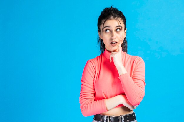 Thoughtful cute girl put her finger to her cheek and thinking on blue background