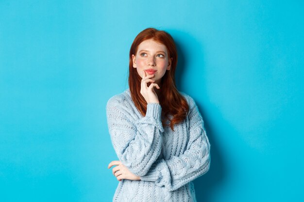 Thoughtful cute woman with red hair, looking upper left corner logo and thinking, imaging something, standing over blue background.