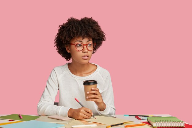 Thoughtful dark skinned girl in round spectacles, makes notes on blank sheet of paper, holds takeaway coffee