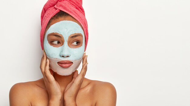 thoughtful dark skinned model applies facial clay mask, wrapped in towel