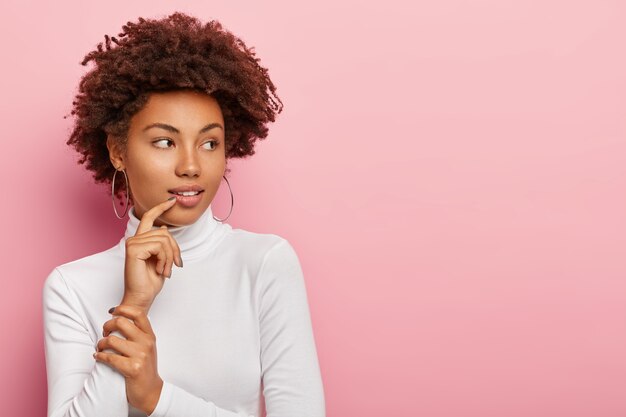 アフロのヘアカットをした思いやりのある暗い肌の女性は、脇を見て、何かについて考え、白いタートルネックのセーター、大きな丸いイヤリングを着て、何かに気づきます