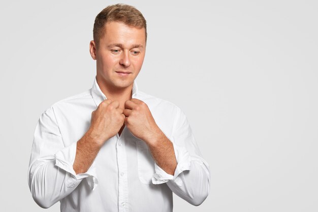 Thoughtful determined businessman in elegant formal white shirt, thinks about something, focused down, has appealing look, stands against white wall with blank copy space for your text