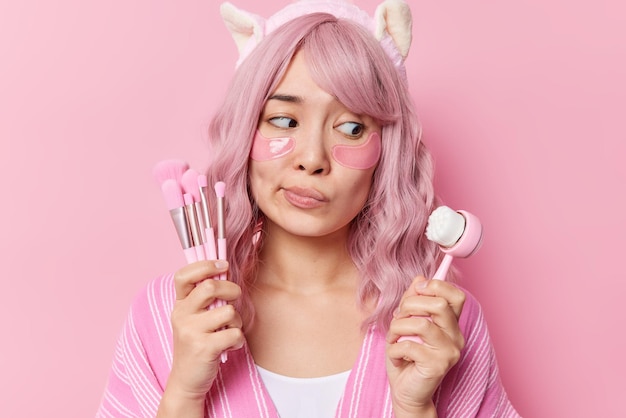 Thoughtful doubtful young woman chooses between face massager and cosmetic brushes undergoes beauty procedures applies hydrogel patches under eyes wears headband isolated over pink background