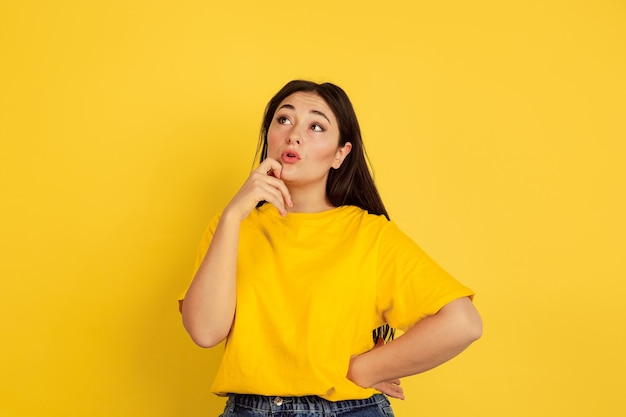 Thoughtful, dreamful. Caucasian woman's portrait isolated on yellow  wall. Beautiful brunette model in casual style. Concept of human emotions, facial expression, sales, copyspace.