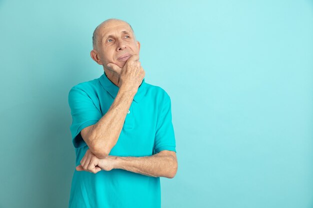 Thoughtful, dreaming. Caucasian senior man's portrait isolated on blue studio background. Beautiful male emotional model. Concept of human emotions, facial expression, sales, wellbeing, ad. Copyspace.