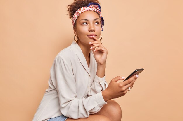 Thoughtful dreamy Afro American woman holds mobile phone in hand