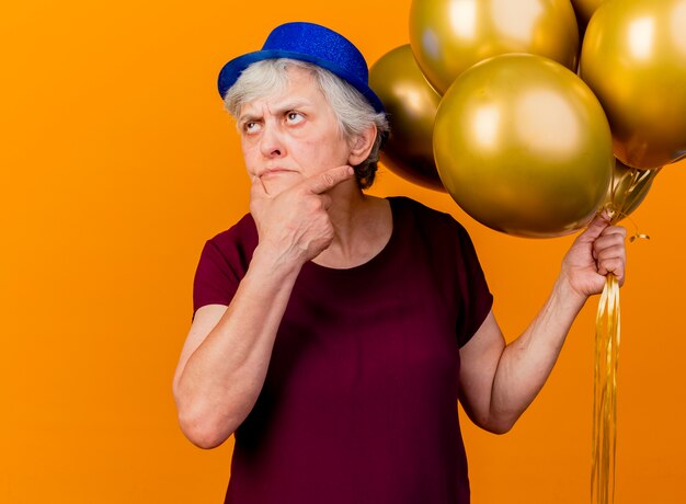 Задумчивая пожилая женщина в праздничной шляпе кладет руку на подбородок и держит гелиевые шары, глядя на оранжевый