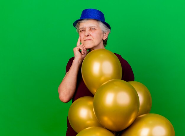 Thoughtful elderly woman wearing party hat stands with helium balloons isolated on green wall with copy space