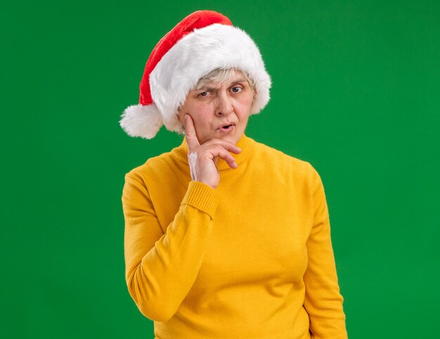 サンタの帽子をかぶった思いやりのある年配の女性は、あごに手を置き、コピースペースで紫色の背景に分離されたカメラを見る