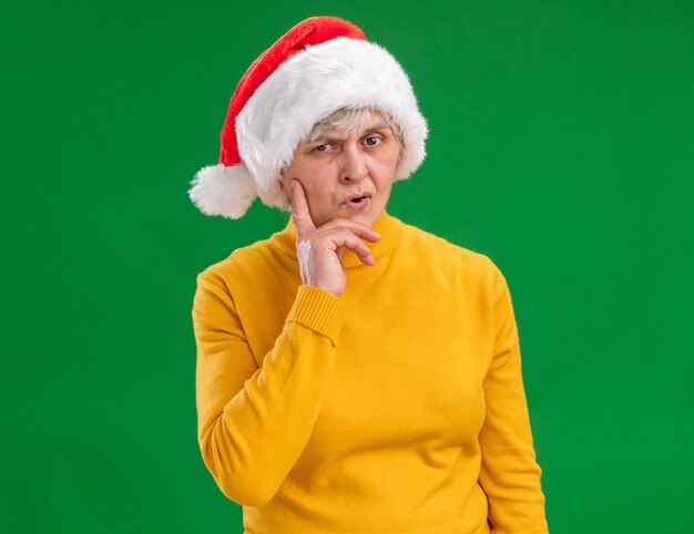 Thoughtful elderly woman with santa hat puts hand on chin and looks at camera isolated on purple background with copy space