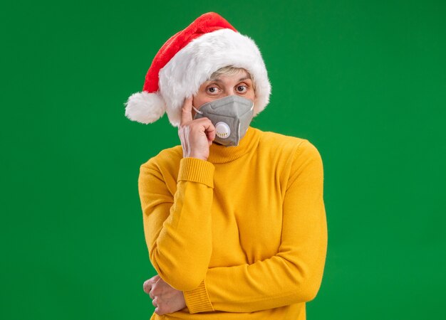 Thoughtful elderly woman with santa hat wearing medical mask puts finger on temple and looks at camera isolated on green background with copy space