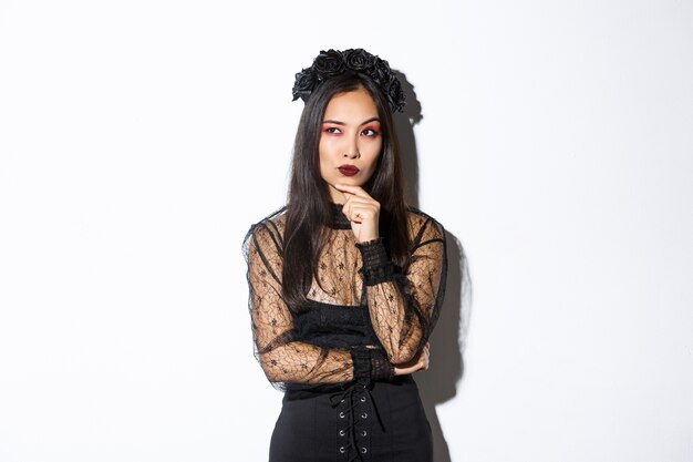 Thoughtful elegant evil witch in black lace dress, gothic wreath and makeup looking upper left corner. Woman making plans for halloween party, thinking over white background.