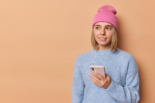 Thoughtful fair haired millennial woman wears pink hat and casual jumper uses mobile phone chats online or browses internet poses against beige background blank space for yoour advertising content