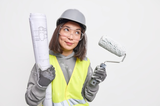 Thoughtful female building contractor looks into distance holds paint roller and blueprint develops ew project wears protective helmet glasses reflective safety vest 