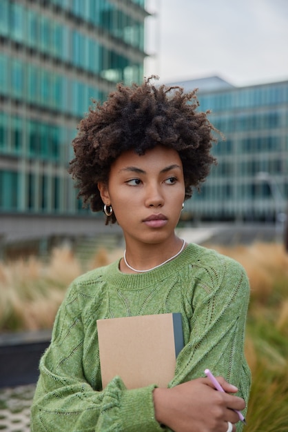 Free photo thoughtful female remote worker holds notepad and pen writes down notes in planner going to make project strolls outside wears green jumper looks away