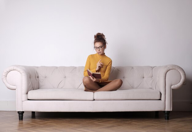 Premurosa donna seduta sul divano e tenendo una matita con un libro sotto le luci