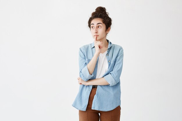 Free photo thoughtful female with hair bun, keeps finger on lips, looks with pensive expression aside, in denim shirt and brown trousers. young woman stares aside while making important decision