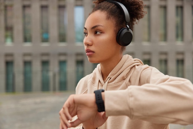 Thoughtful fitness woman checks results of cardio training on smartwatch focused away wears casual hoodie listens music via headphones poses outdoors near building. Sporty lifestyle concept.