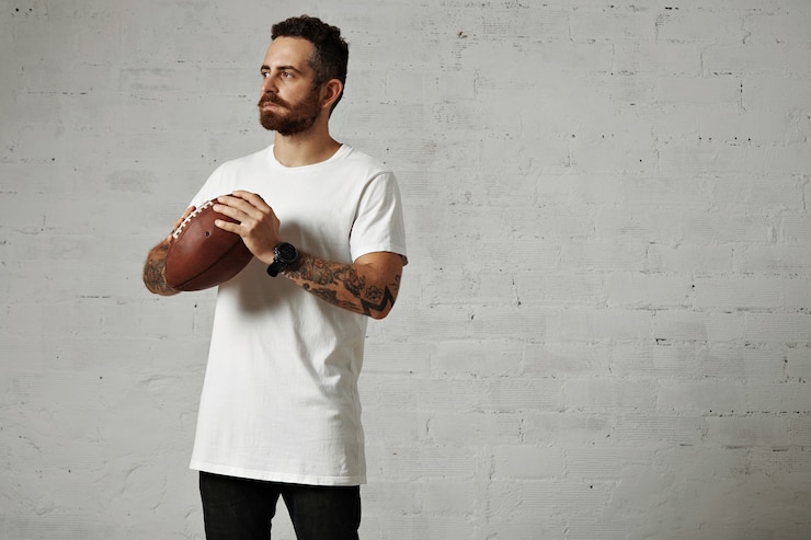 Free Photo thoughtful focused athletic stylish young man with tattoos and beard holding a vintage leather rugby ball on white wall