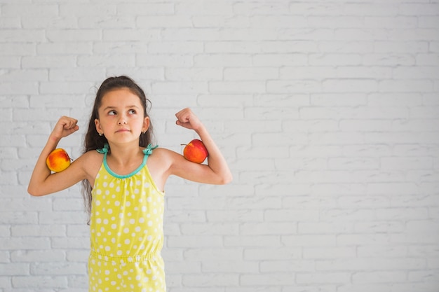 Thoughtful girl standing against white wall holding apples on her biceps